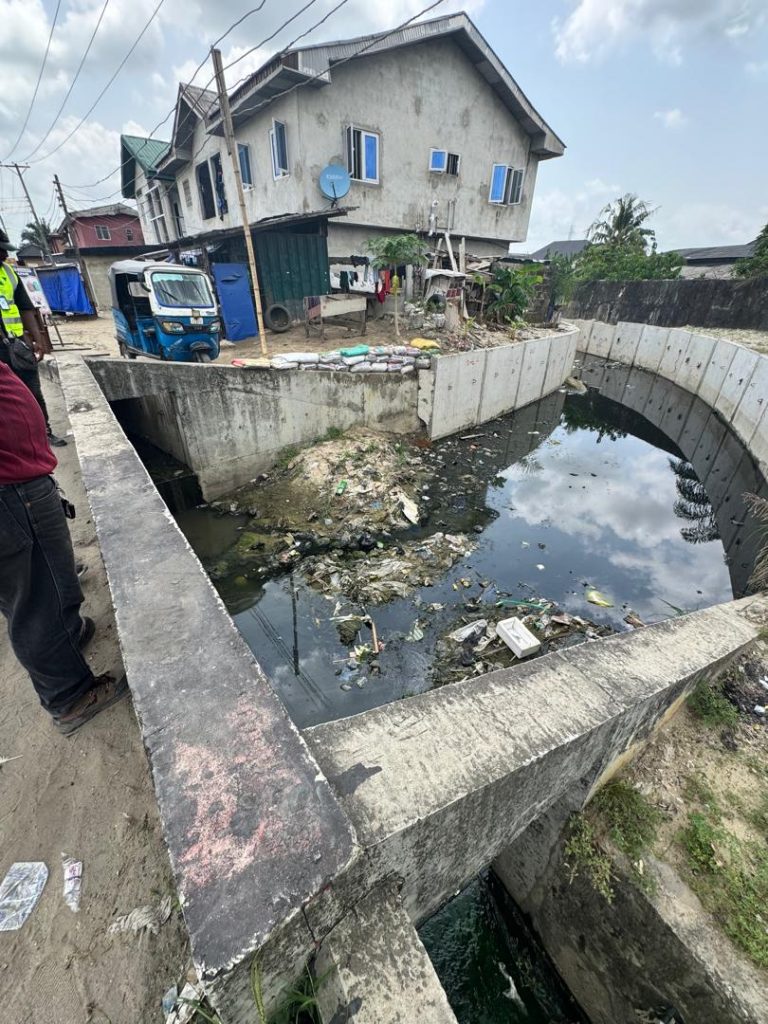 Curved concrete drainage channel section showing waste accumulation reducing effective flow area. Stagnant water conditions indicate complete hydraulic failure in this segment. The curved geometry, designed for flow transition, has been compromised by indiscriminate waste disposal.